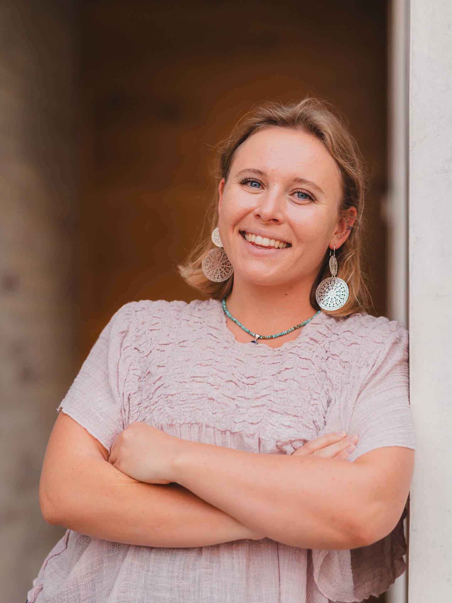 Ainsley Bost, smiling, arms crossed, leaning against a doorway in natural light