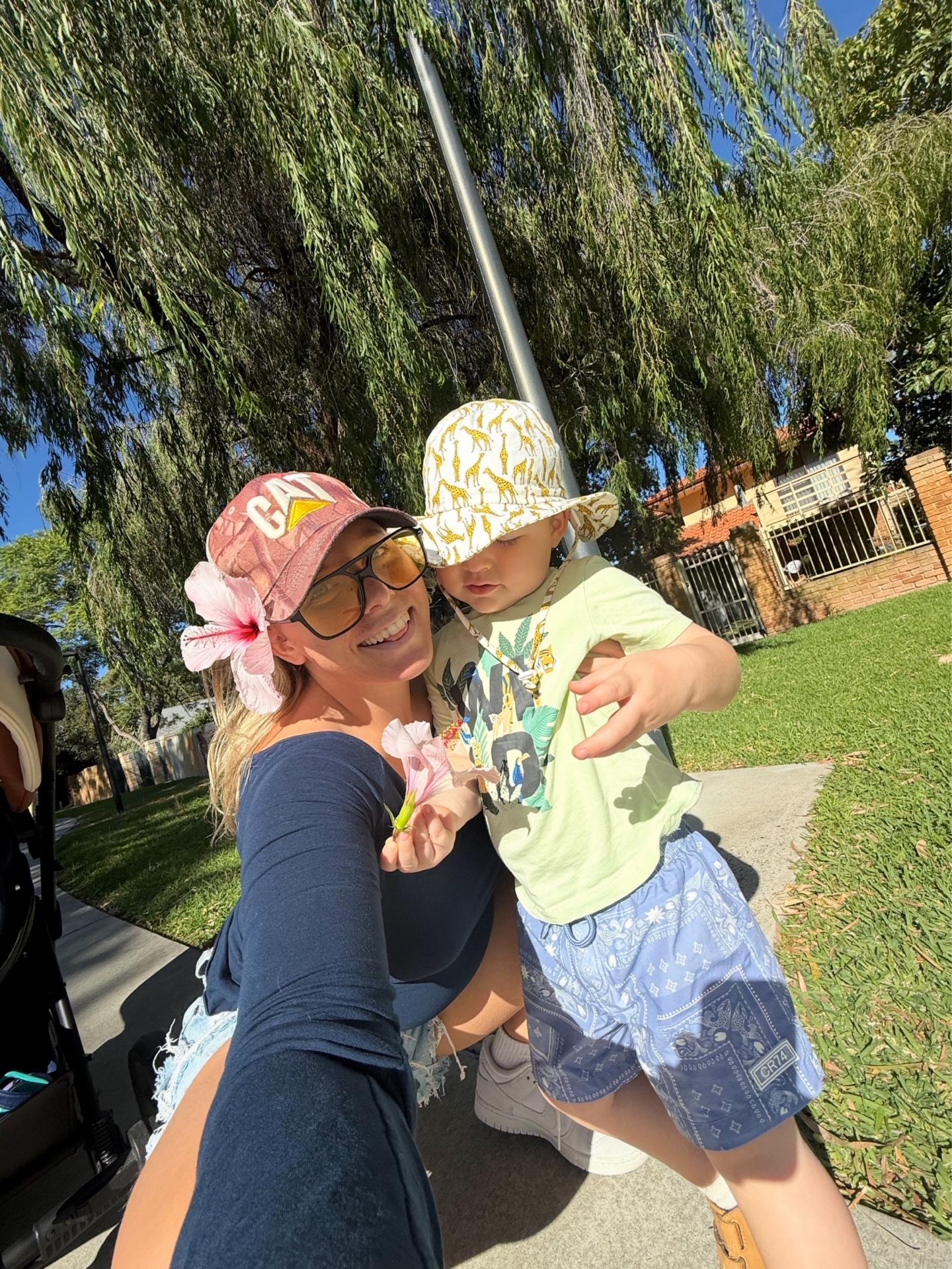 Ainsley smiling in the sunshine with a toddler in a giraffe-print sun hat, a leafy park behind them